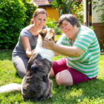 Two smiling women sitting on the grass petting a dog
